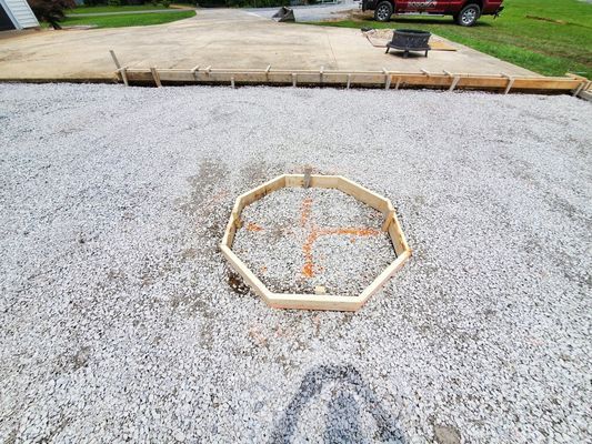 An octagon-shaped wooden frame sits on a gravel driveway, with orange markings on the gravel indicating a placement point.