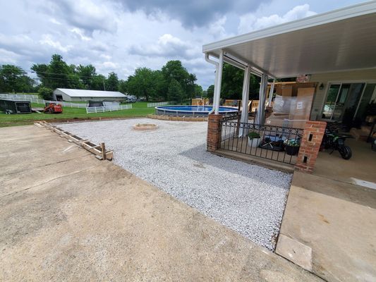 A backyard patio area under construction, featuring a fresh gravel surface, wooden forms, and a pool in the background.