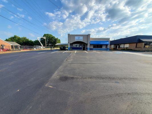 A low-angle view of a grey commercial building with blue trim, seen from a large, freshly paved asphalt parking lot.