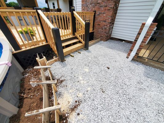 A wooden staircase leads from a deck to a gravel patio area under construction, with wooden forms set up nearby.