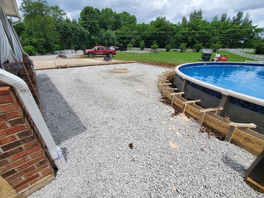 A gravel patio under construction next to a round above-ground swimming pool with wooden forms installed around its base.