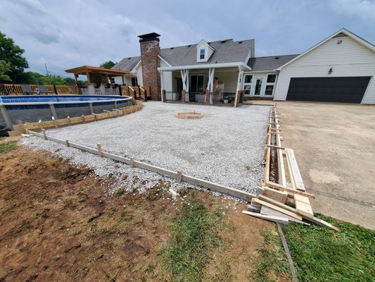 A gravel-filled backyard area with wooden forms set up for a new concrete patio, next to a pool and a house.