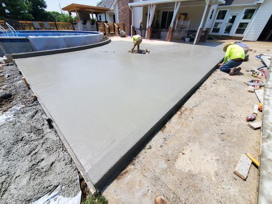 Workers in high-visibility vests finish a freshly poured concrete patio next to an above-ground pool and house patio.