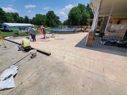 Workers applying a decorative finish to a freshly poured concrete patio near an outdoor pool.