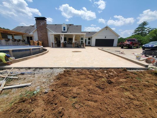 A wide, light-colored concrete patio adjacent to a swimming pool and house, with freshly turned dirt in the foreground.