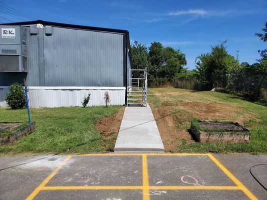 A concrete sidewalk leads to a metal stair set at the side entrance of a grey industrial building on a sunny day.