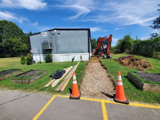 An orange excavator parked next to a gravel path leading to a metal building, marked by two traffic cones.
