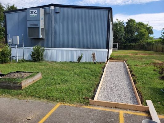 A gravel walkway bordered by wooden frames leads from a paved area toward a blue metal building with an attached unit.