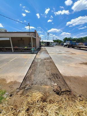 A long, narrow trench cut through a concrete parking lot, showing exposed gravel and dirt, with a building to the left.