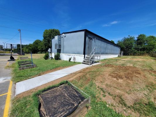 A gray modular building with a concrete walkway and stairs, set in a grassy area with a basketball hoop nearby.