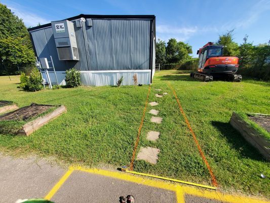 An orange excavator sits on a grassy lawn next to a corrugated metal building, with a marked path for measurement.