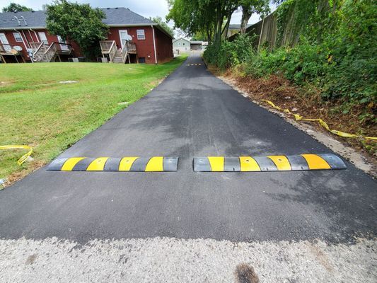 A freshly paved asphalt driveway with two black and yellow speed bumps installed across the width of the path.