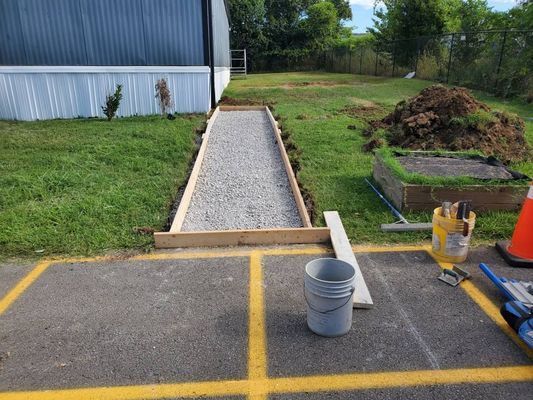 Wooden frame with gravel base for a walkway near a building, with a bucket and tools nearby on asphalt.