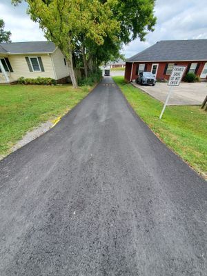 A newly paved asphalt driveway leads between a yellow house and a red brick building with a 