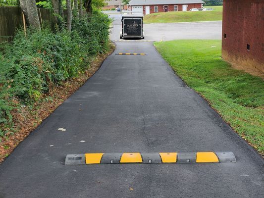 Two black and yellow speed bumps on a paved driveway, with a trailer parked in the background.