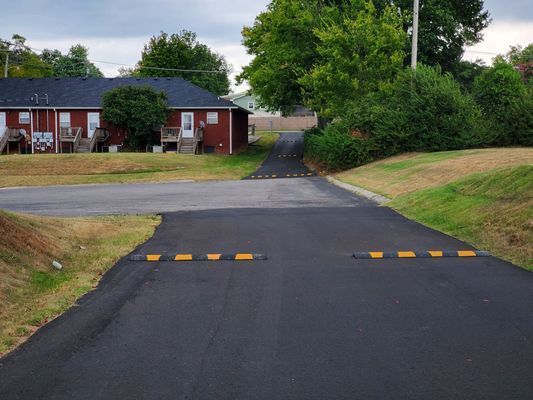 A paved driveway featuring two black and yellow speed bumps leads toward a red residential building under a cloudy sky.