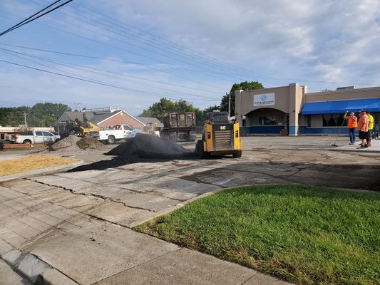 A yellow skid-steer loader works on a construction site in a commercial parking lot, with workers standing nearby.