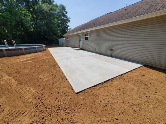 A freshly poured concrete patio sits next to a house with beige siding, adjacent to an above-ground pool and dirt yard.