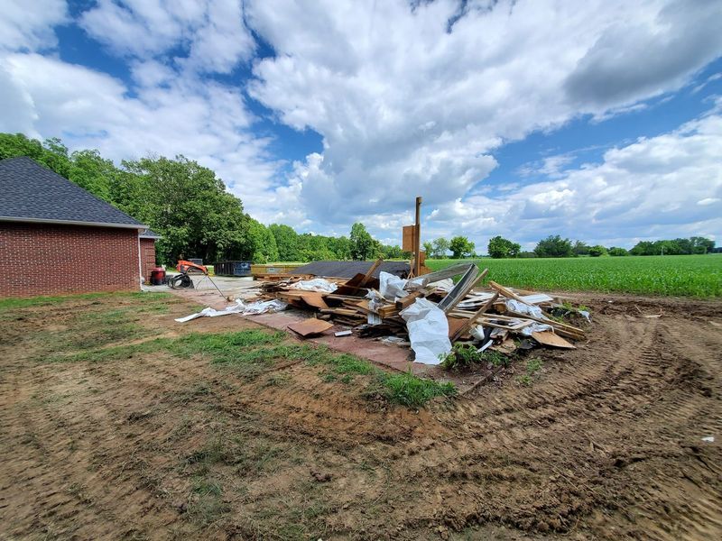 A brick house exterior next to a debris pile from a demolition project on a dirt lot, with a field and blue sky beyond.
