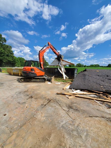 An orange excavator lifts a piece of building debris into a large dumpster on a sunny, grassy work site.