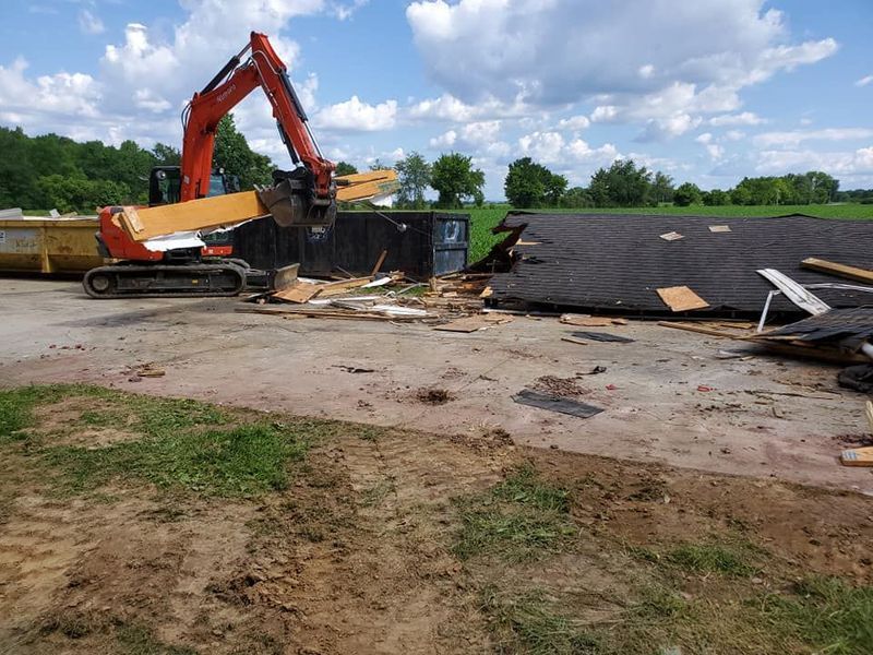 An orange excavator lifts a wooden beam near a collapsed building and a dumpster on a sunny, grassy property.