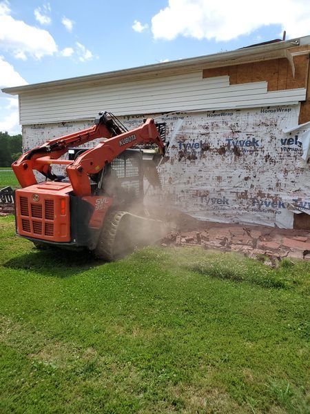 An orange skid-steer loader clears debris from a building exterior covered in Tyvek wrap and partial wood siding.
