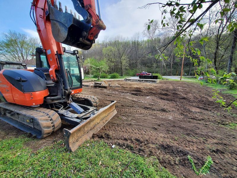 An orange excavator sits on a dirt lot undergoing site preparation with a forest background.