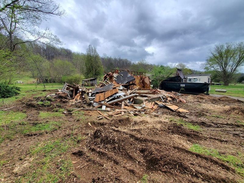 A debris pile from a demolished building in a grassy field with a large black dumpster nearby under a cloudy sky.