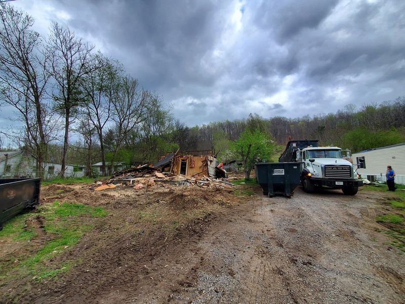 A demolished building site with piles of debris, a large dumpster, and a dump truck under a cloudy sky.