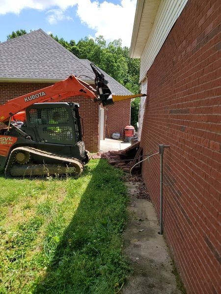 An orange skid steer loader operates near the brick exterior wall of a house, positioned in a yard with green grass.