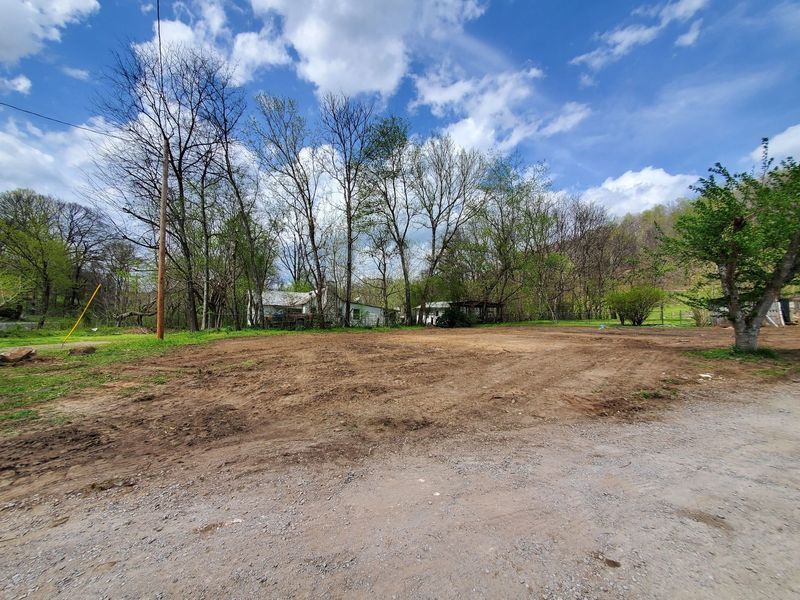 A cleared, dirt-covered lot in a rural setting, bordered by trees and a blue, cloudy sky.