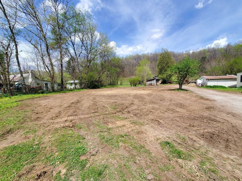 A cleared, dirt-covered lot on a sunny day with trees and houses in the background and a gravel road on the right.