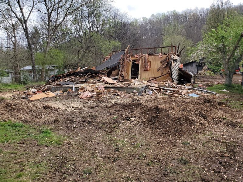 A demolished house with scattered debris on a dirt lot, surrounded by trees and a clear sky.