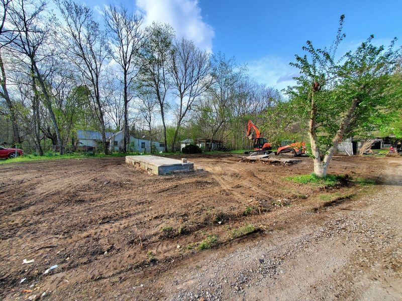 A cleared dirt lot features a concrete foundation slab with an orange excavator in the background under a blue sky.