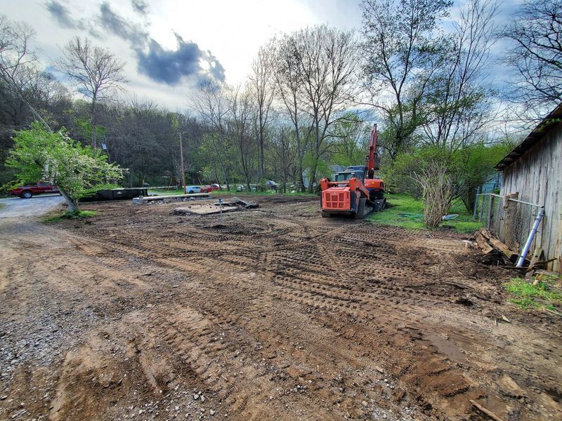 An orange excavator parked on a muddy, cleared plot of land near trees and a rustic shed under a cloudy sky.