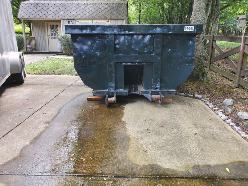 A dark green, empty construction dumpster sits on a concrete driveway near a storage shed and a wooden fence.