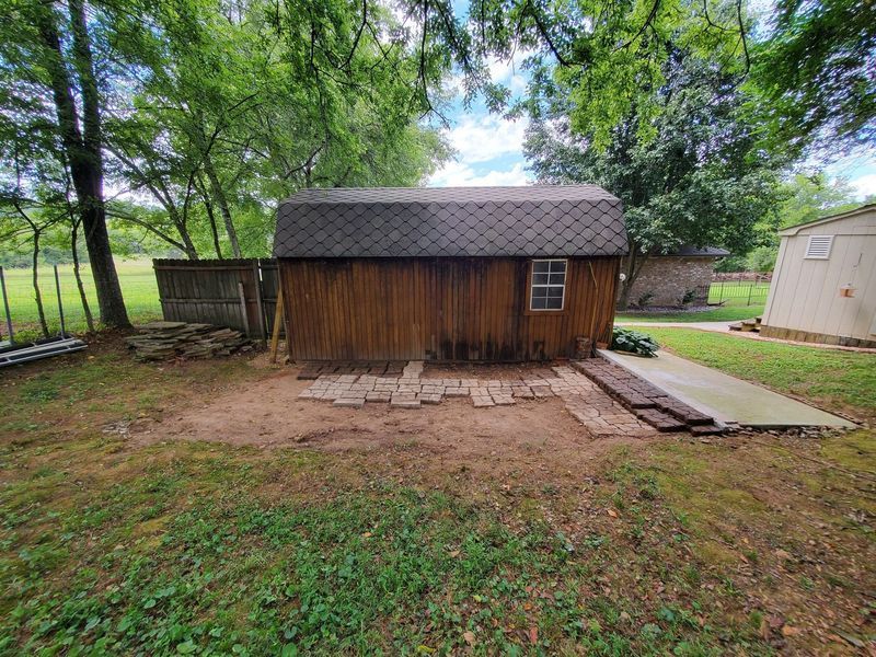 A rustic wooden storage shed in a grassy backyard with a partially paved stone walkway leading up to it.