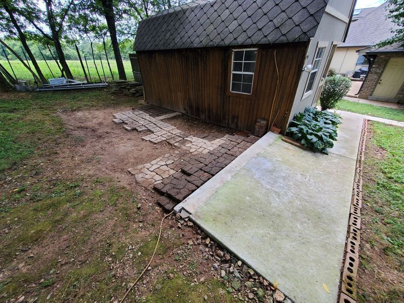 A backyard shed next to a concrete patio with a work-in-progress brick pathway being laid on the adjacent bare soil.