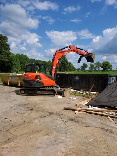 An orange excavator parked in a lot, lifting a claw over a large dark dumpster filled with construction debris.