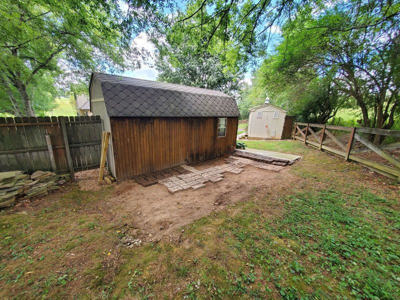 A wooden shed with a dark roof sits in a backyard next to a fence, with a partial brick walkway laid on the ground.
