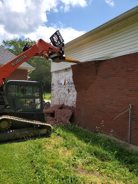 An orange Kubota skid steer uses its front attachment to peel a section of brick siding off a residential house exterior.