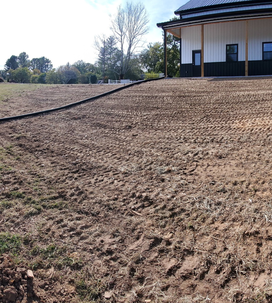 A modern white house with a dark metal roof sits on a sloped, recently cleared plot of dirt with a landscape border.