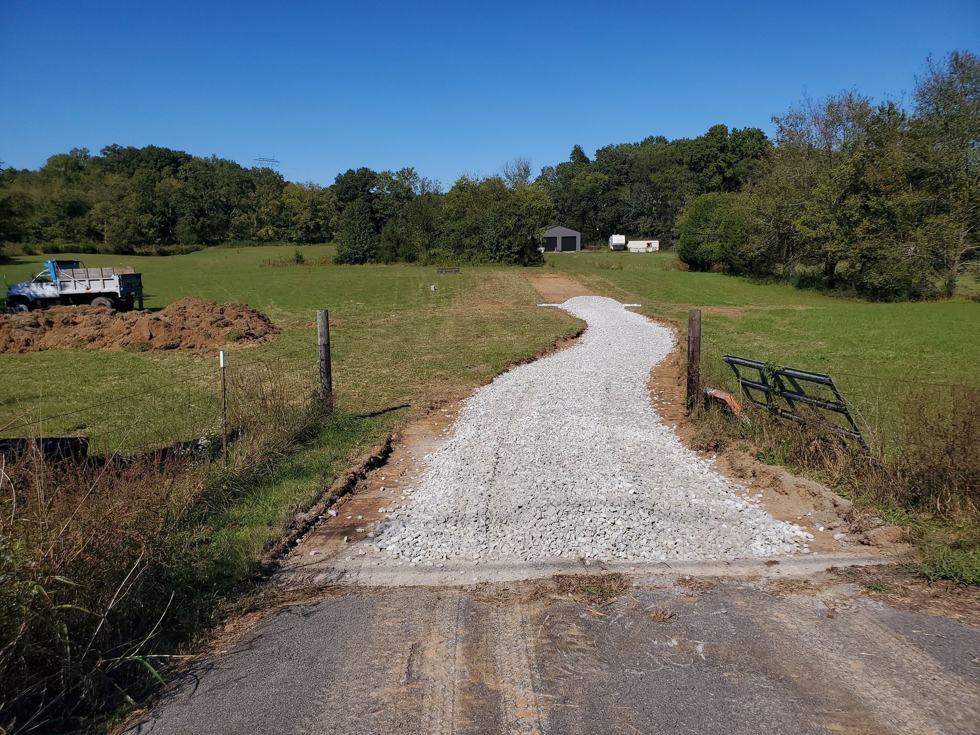 A gravel driveway leads from an asphalt road into a green field with trees and a truck visible in the distance.