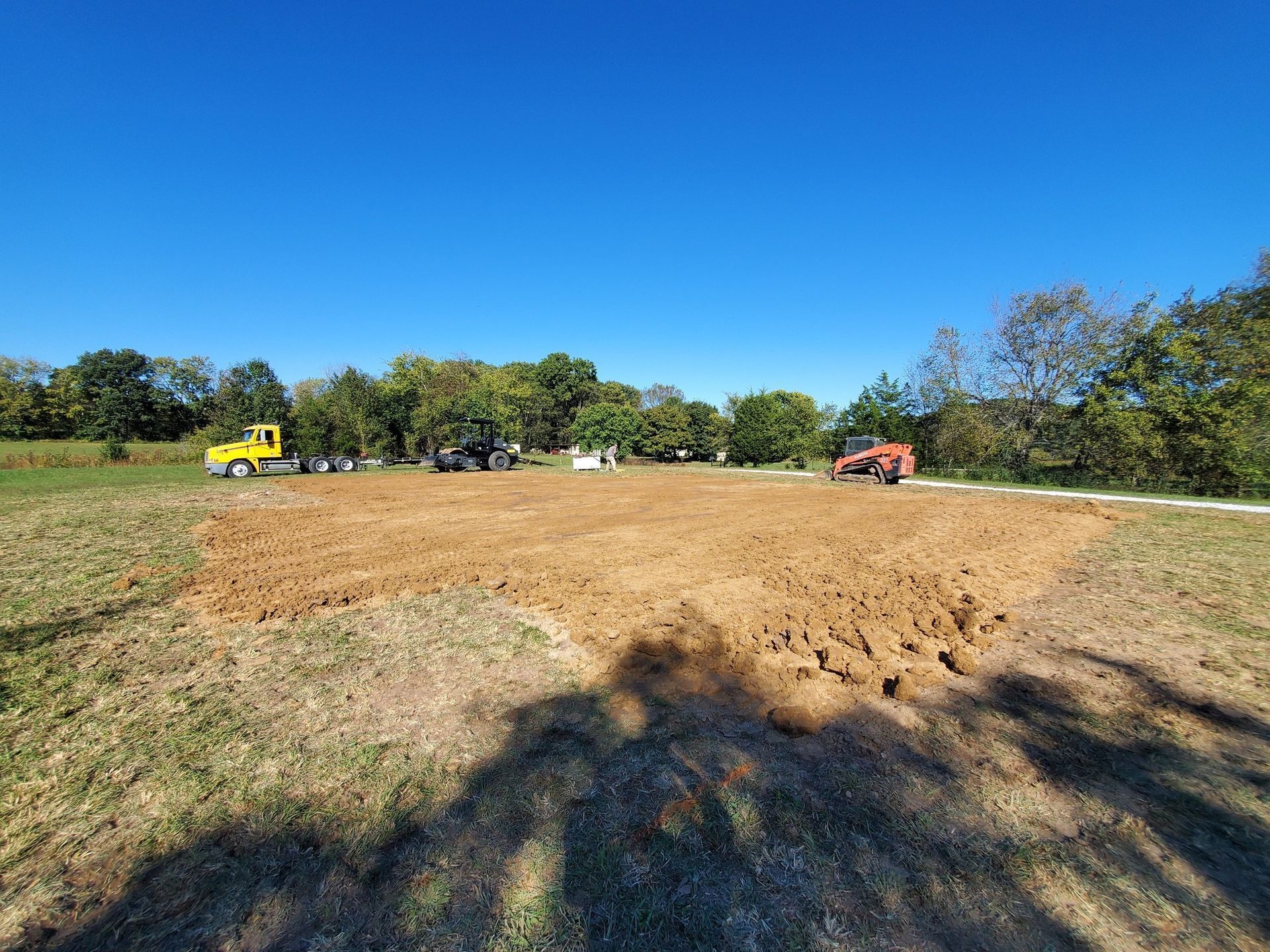A yellow truck and a red tractor work on a large, freshly tilled brown soil patch in a grassy field under a blue sky.