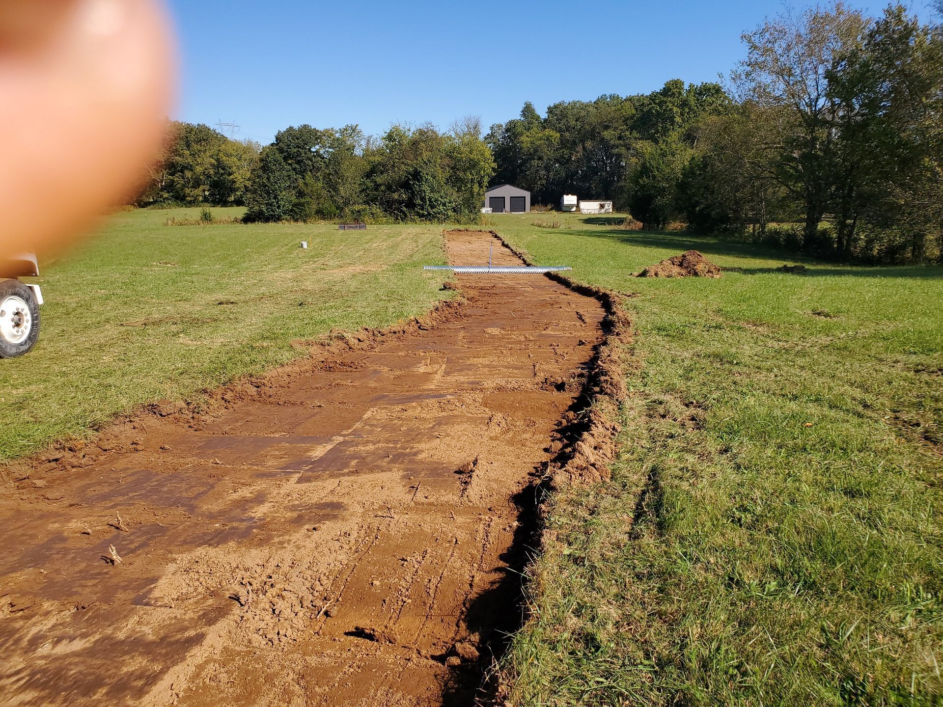 A dirt trench cuts through a grassy field toward a distant building, with part of a vehicle visible on the left.