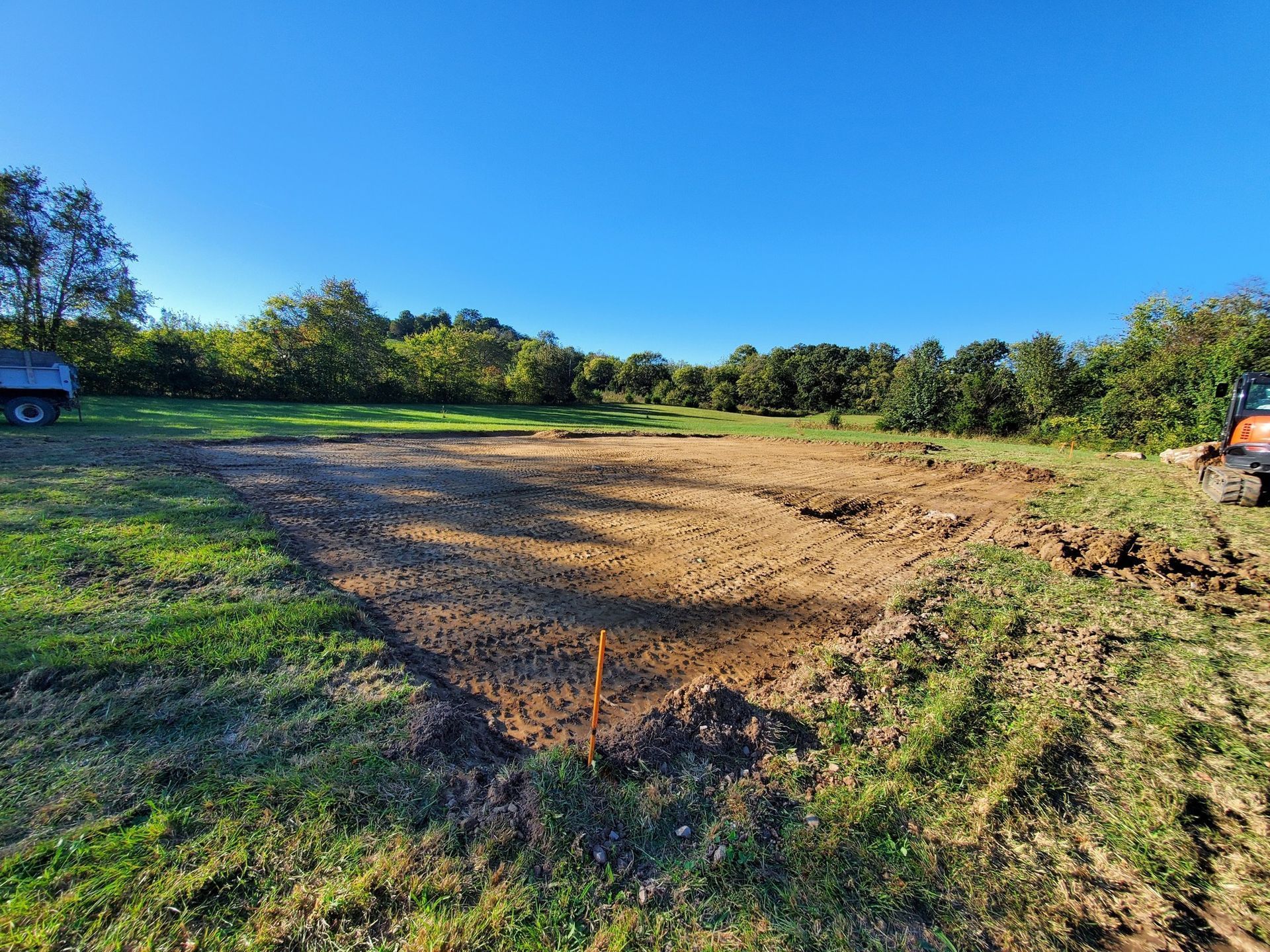 An excavated rectangular plot of bare dirt in a grassy field under a clear blue sky, with trees in the background.