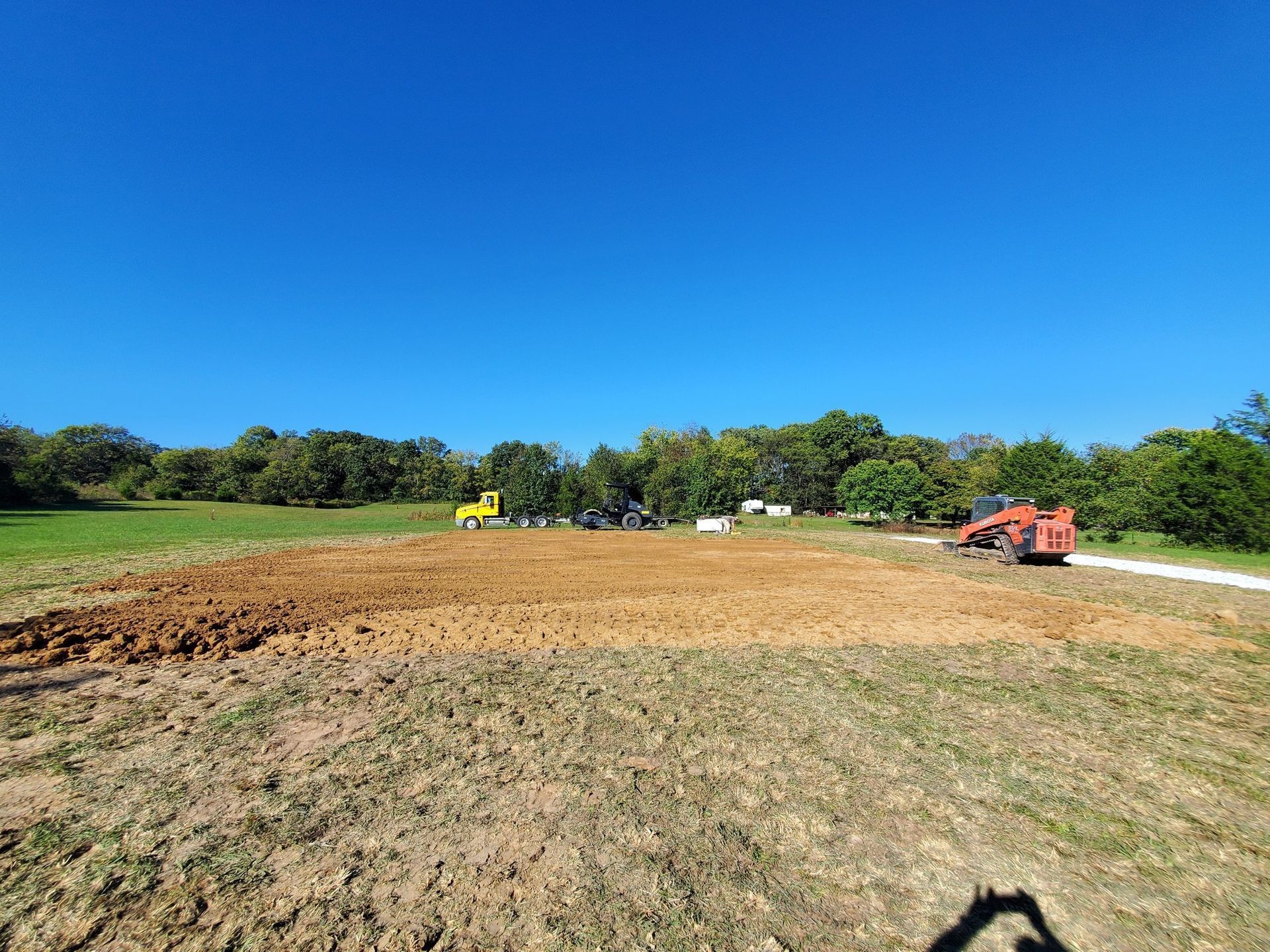 A construction site in a field with a yellow truck, a red tractor, and cleared brown earth under a clear blue sky.