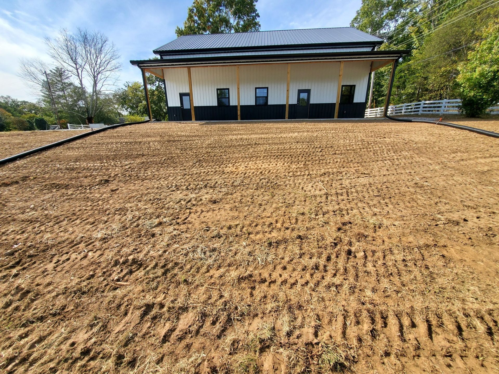A white building with a dark roof and foundation sits behind a freshly graded, dirt yard with visible tire tracks.