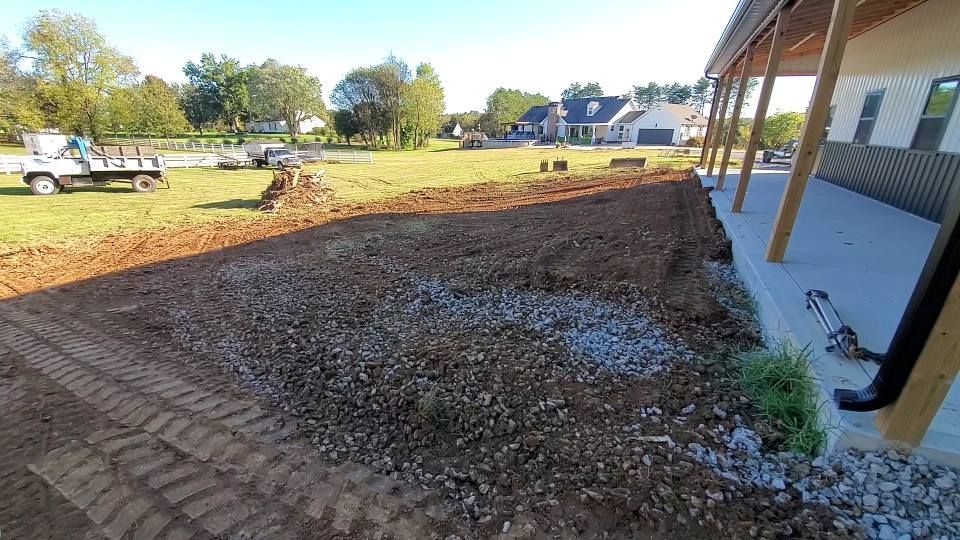 A wide, dirt-covered yard next to a concrete porch with wooden support posts, with a truck parked in the grassy distance.