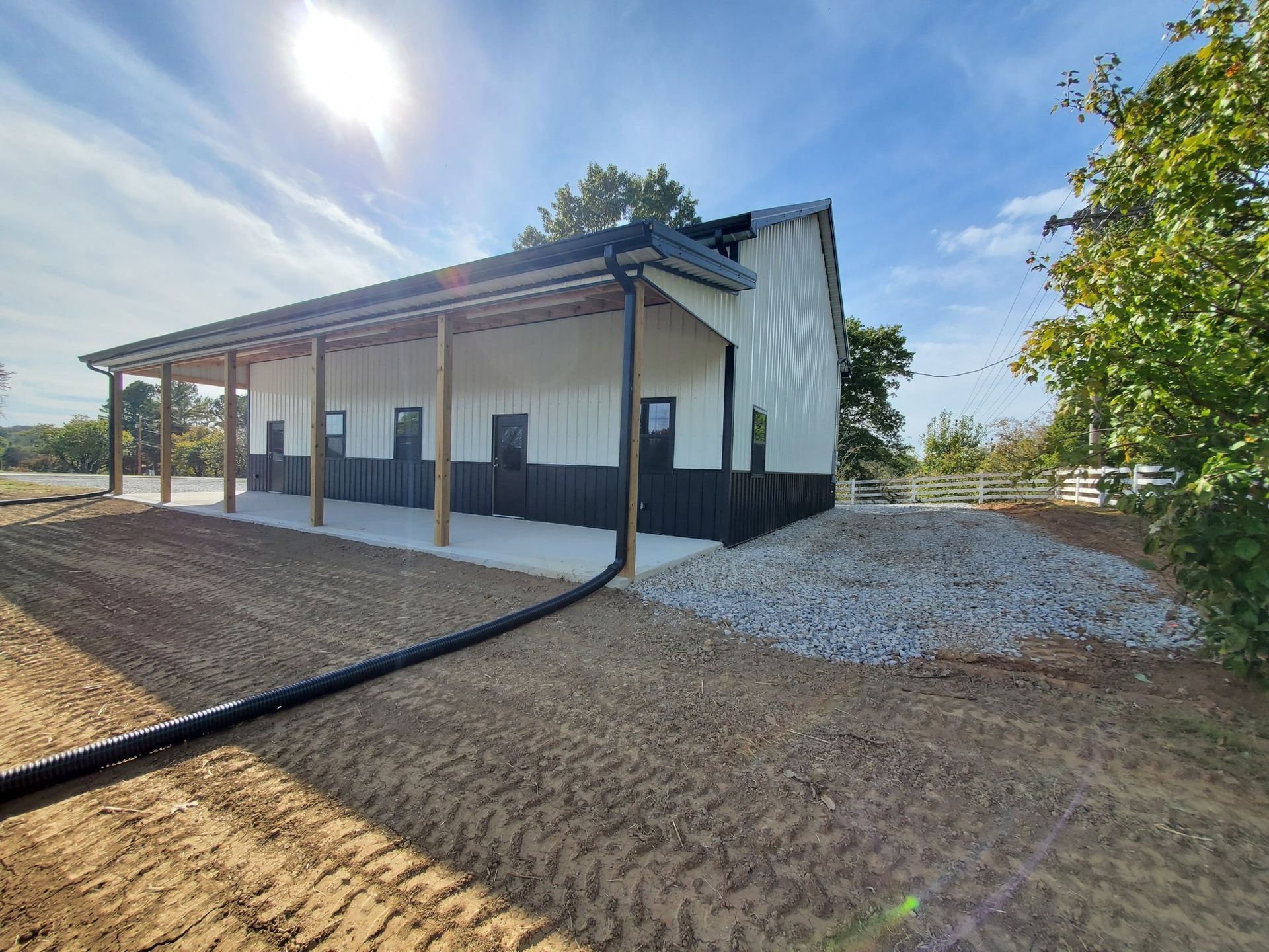 A modern white and black metal building with a covered porch sits on a gravel lot under a sunny blue sky.